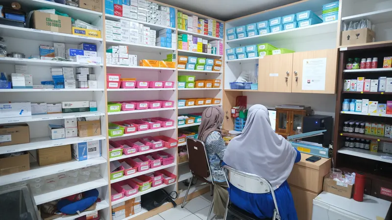 Instalasi farmasi Klinik Pratama Waluya Sentosa, rak obat tertata dalam wadah berlabel dari lantai sampai bagian atas, dengan dua petugas sedang bekerja di meja di sudut ruangan.