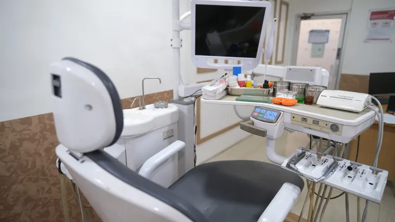Ruang praktik dokter gigi dengan kursi periksa gigi (dental chair), lampu tindakan, dan meja alat berisi perlengkapan pemeriksaan gigi.
